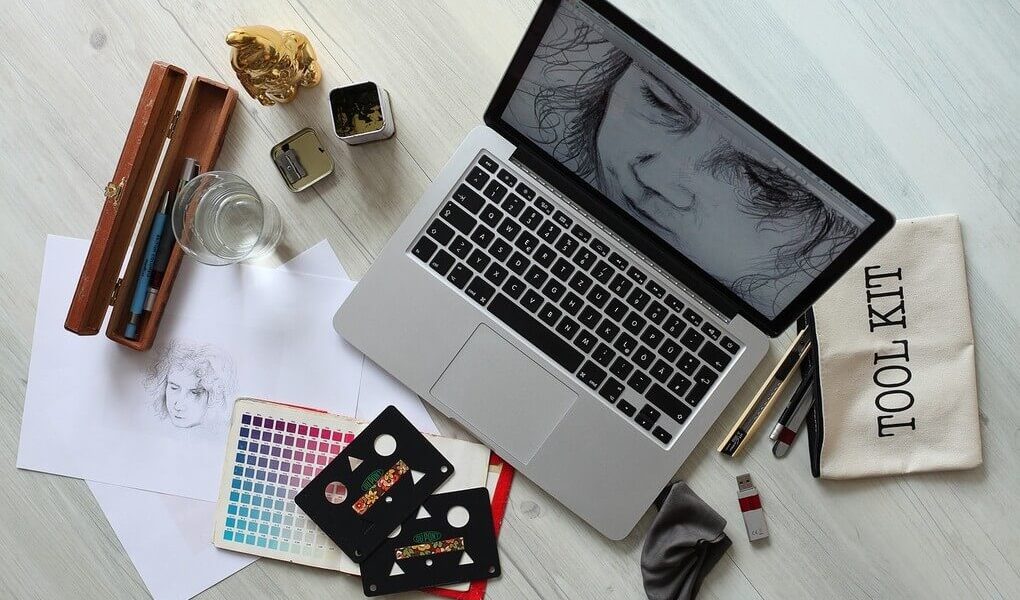 A wooden table displaying a laptop, sketchbook, and various drawing materials in a creative workspace.