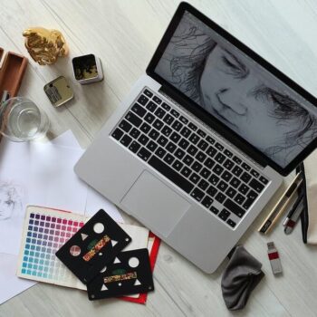 A wooden table displaying a laptop, sketchbook, and various drawing materials in a creative workspace.