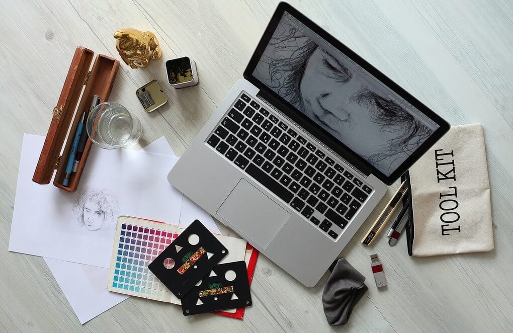 A wooden table displaying a laptop, sketchbook, and various drawing materials in a creative workspace.