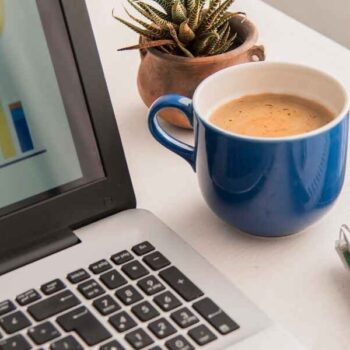 A laptop displaying colorful charts, next to a blue coffee mug, small catus plant, a tablet, and a notebook with a pen. A productive and focused workspace.