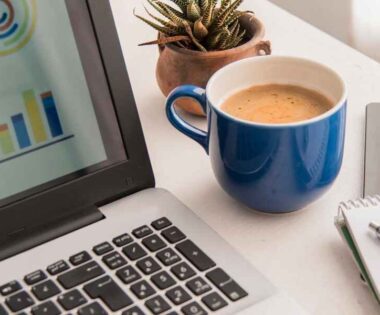 A laptop displaying colorful charts, next to a blue coffee mug, small catus plant, a tablet, and a notebook with a pen. A productive and focused workspace.