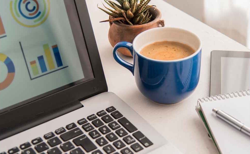 A laptop displaying colorful charts, next to a blue coffee mug, small catus plant, a tablet, and a notebook with a pen. A productive and focused workspace.