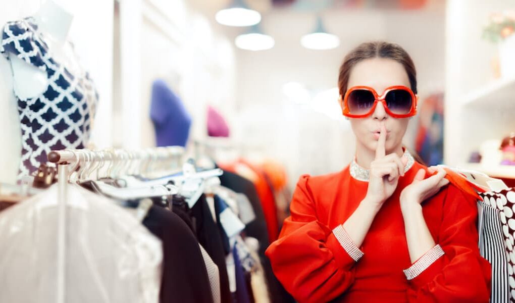 A woman in a vibrant red dress, wearing sunglasses, holding hangers with clothes, in a clothes shop.