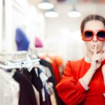 A woman in a vibrant red dress, wearing sunglasses, holding hangers with clothes, in a clothes shop.