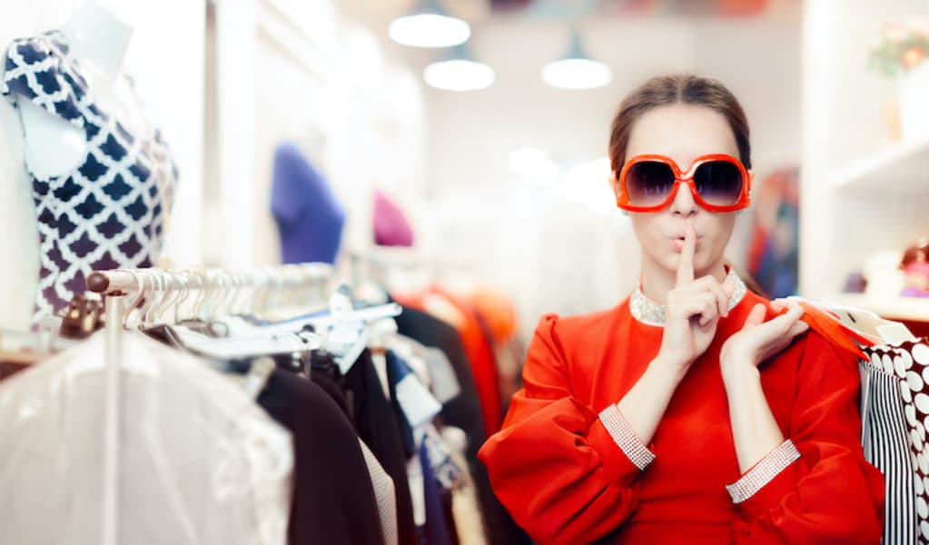 A woman in a vibrant red dress, wearing sunglasses, holding hangers with clothes, in a clothes shop.