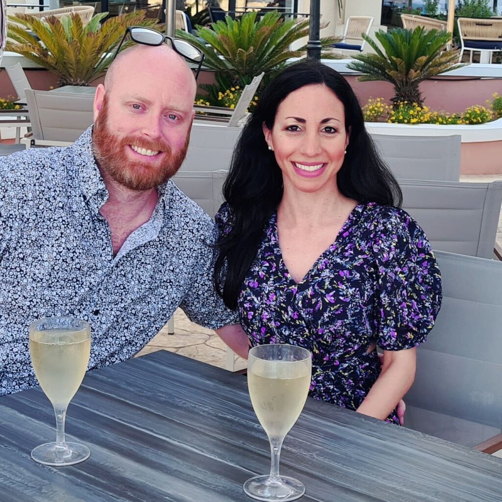 A smiling couple sits at a patio table with two glasses of white wine. The setting is relaxed, with palm plants and neutral-toned decor in the background.