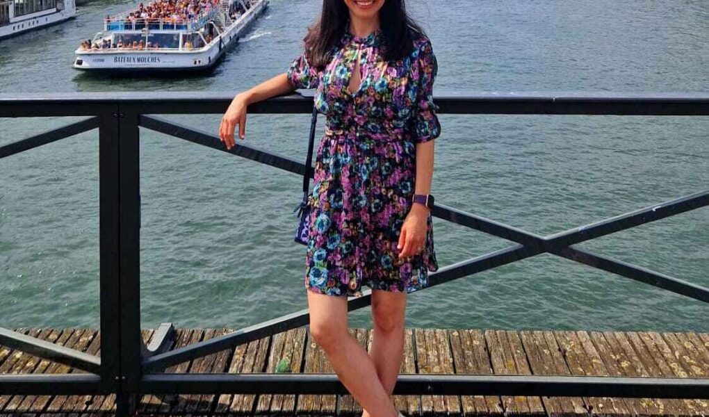 A woman in a brightly coloured floral dress leans on a wooden railing by a river, smiling. A tour boat filled with people sails past, with trees lining the riverbank.