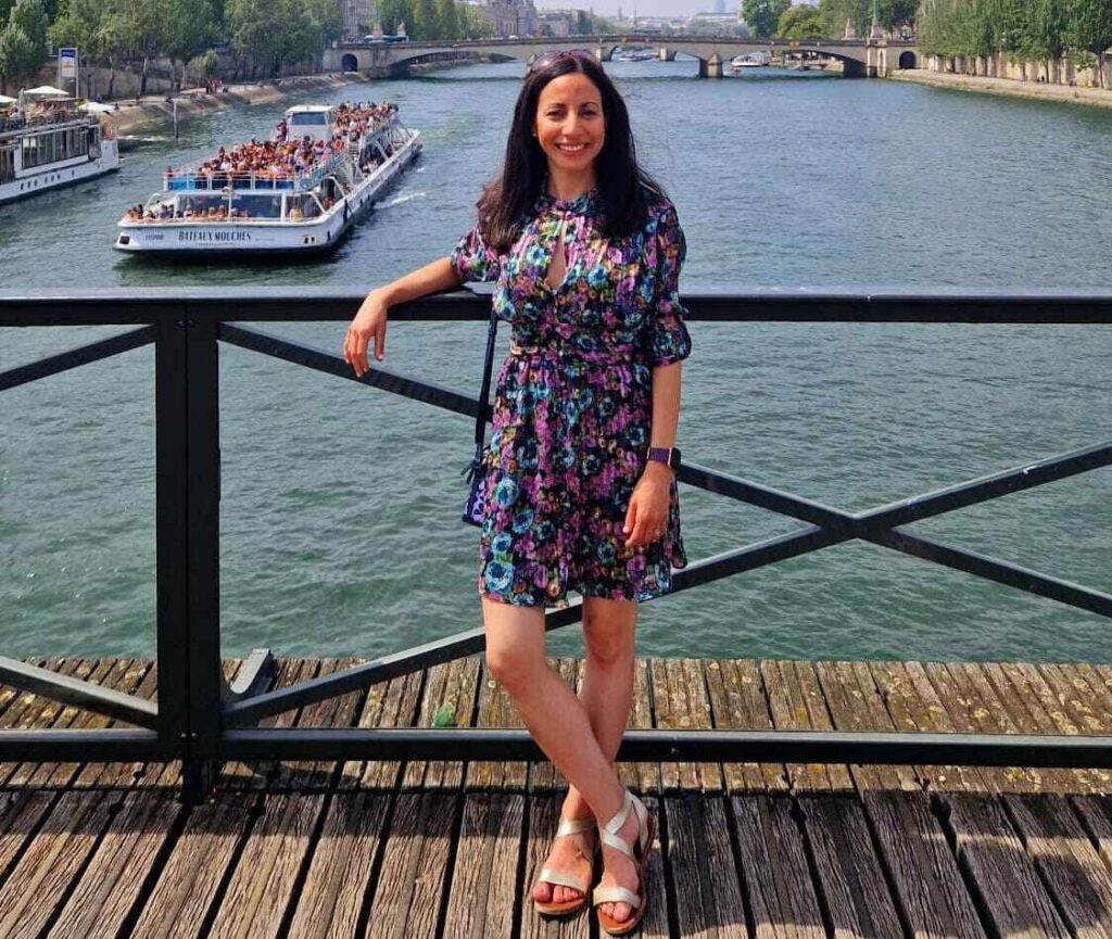 A woman in a brightly coloured floral dress leans on a wooden railing by a river, smiling. A tour boat filled with people sails past, with trees lining the riverbank.