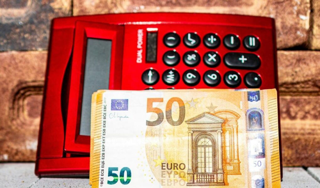 Red calculator with black buttons sits against a brick wall. A 50 Euro banknote is placed in front of it.