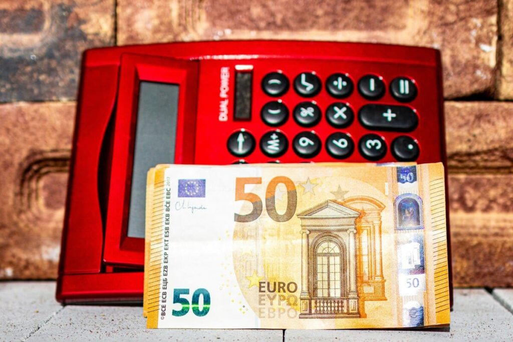 Red calculator with black buttons sits against a brick wall. A 50 Euro banknote is placed in front of it. 