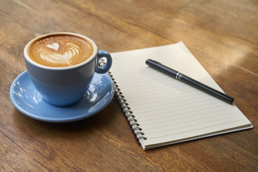 A cappuccino with heart-shaped foam art, next to an open spiral notebook and pen on a wooden table, suggesting a cozy, productive atmosphere.