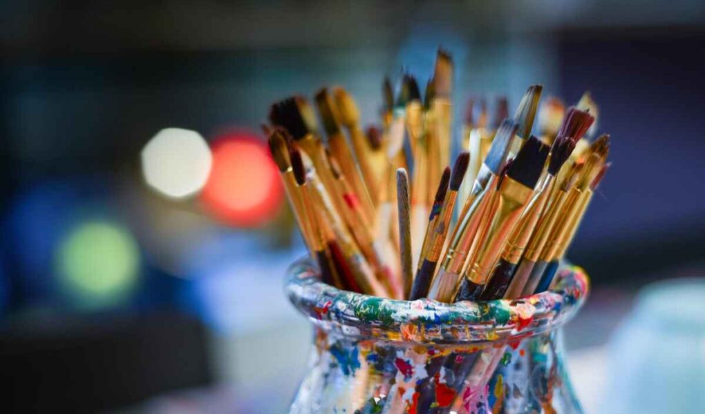 A jar filled with assorted paintbrushes, each varying in size and shape, with colorful paint splatters on the jar. The background is softly blurred.