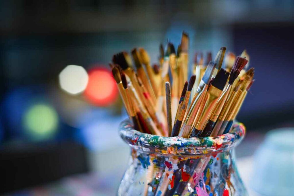 A jar filled with assorted paintbrushes, each varying in size and shape, with colorful paint splatters on the jar. The background is softly blurred.