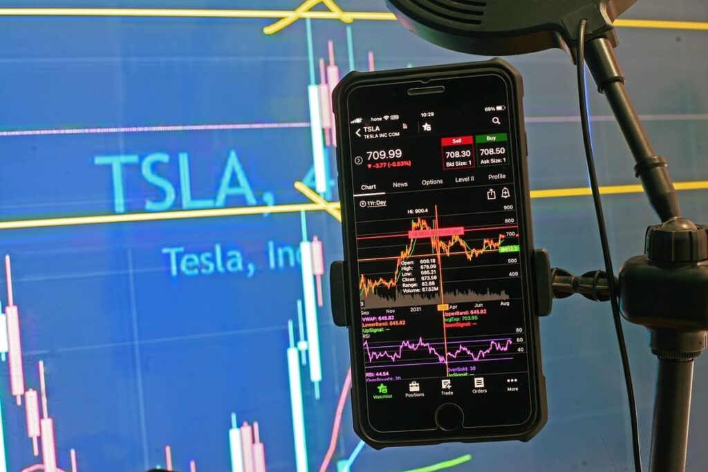 Smartphone displaying Tesla stock chart with detailed analytics, in front of a large screen showing Tesla's name and financial data. High-tech, analytical tone.