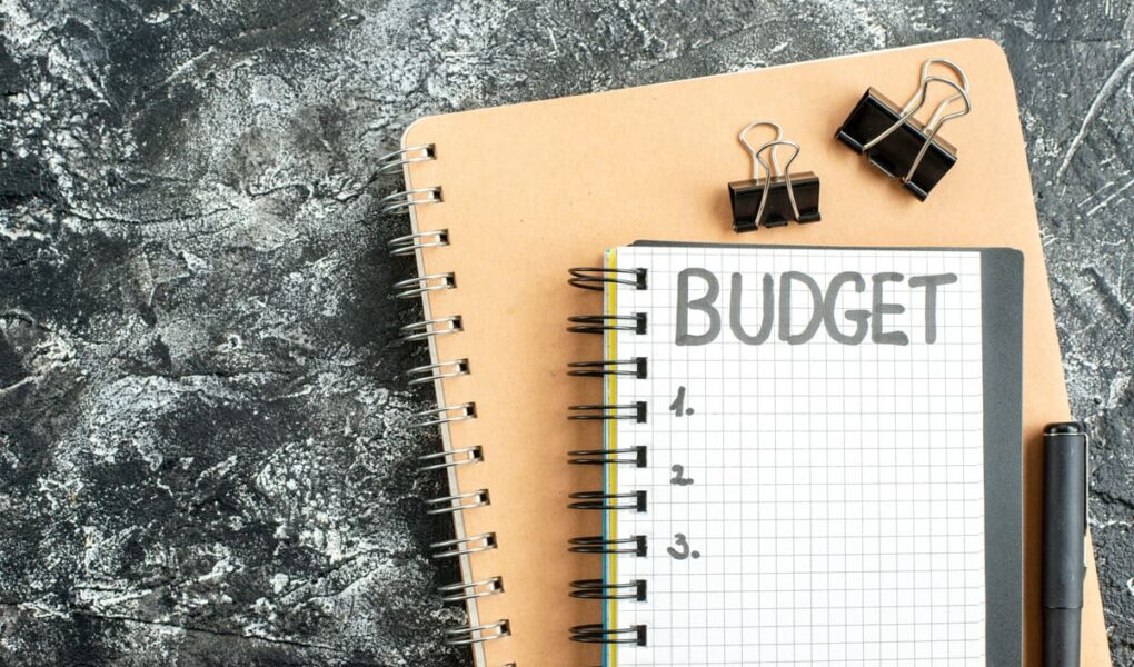 A notebook with the word ‘budget’ sits on a larger brown spiral notepad. Nearby are binder clips and a pen on a textured grey surface.