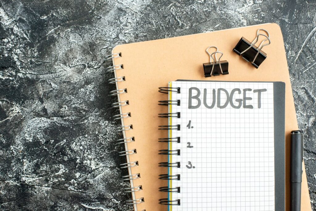 A notebook with the word ‘budget’ sits on a larger brown spiral notepad. Nearby are binder clips and a pen on a textured grey surface.