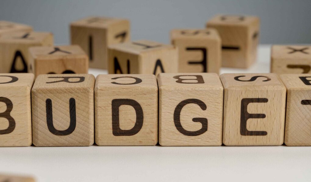 Wooden blocks spelling "BUDGET" are lined up on a white surface, surrounded by scattered, blurred blocks. The tone is focused and organised.